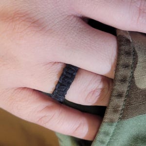 May include: A black ring constructed from woven cord material featuring a textured, knotted pattern that wraps around the finger. The band has a consistent width and a matte finish, showcasing a simple, braided design made from dark fiber strands.