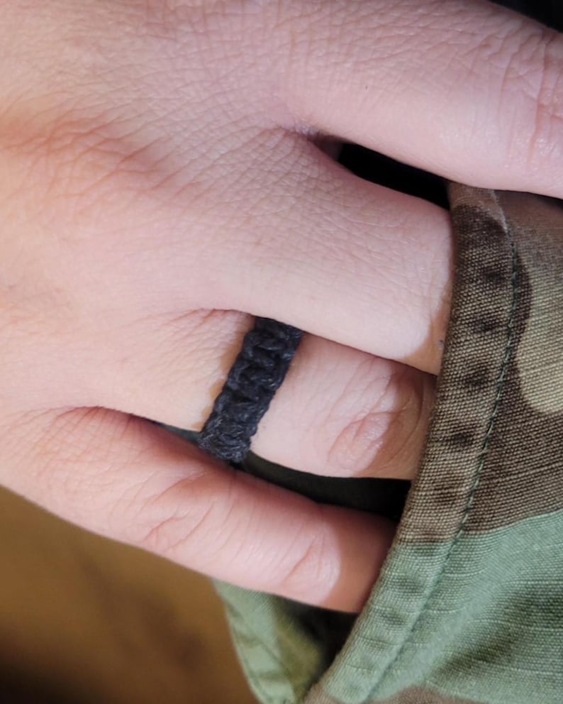 May include: A black ring constructed from woven cord material featuring a textured, knotted pattern that wraps around the finger. The band has a consistent width and a matte finish, showcasing a simple, braided design made from dark fiber strands.