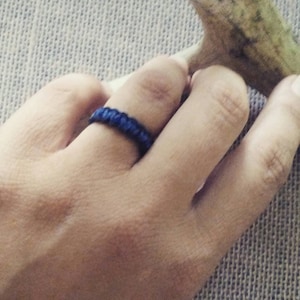 May include: A dark blue woven ring is worn on a finger. The ring is made of a tightly woven material, and the color is a deep, saturated blue. The ring is a simple, yet stylish accessory.