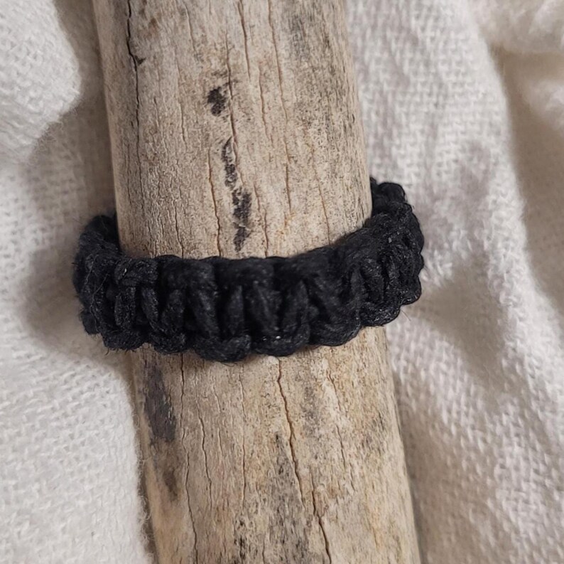 May include: A black ring featuring a braided or knotted cord construction. The band is crafted from a continuous piece of dark, textured material with a repeating woven pattern that creates a uniform, circular shape around the center.