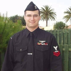 May include: A black military-style uniform featuring a long-sleeved button-down shirt with a matching tie, a silver tie bar, and a black garrison cap. The shirt displays a silver wing insignia, a multi-colored ribbon rack, and a red chevron patch.