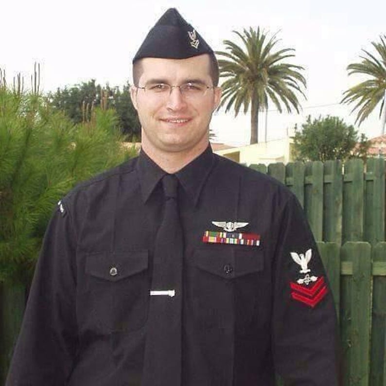May include: A black military-style uniform featuring a long-sleeved button-down shirt with a matching tie, a silver tie bar, and a black garrison cap. The shirt displays a silver wing insignia, a multi-colored ribbon rack, and a red chevron patch.