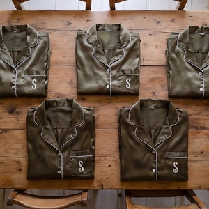 May include: Five olive green satin pajama tops with white piping and a pocket with the letter "S" embroidered on the pocket. The pajamas are arranged on a wooden table, with two wooden chairs visible in the background.