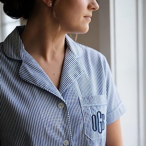 May include: A blue and white striped pajama shirt with a monogrammed pocket. The short-sleeved shirt has a classic collar and button-down front. The person is looking out a window.