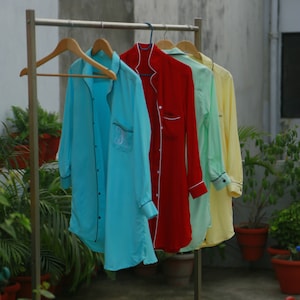 May include: Four long-sleeved sleep shirts in various colors hang on a metal rack. The shirts are light blue, red, light green, and yellow. Each shirt has black trim and a button-down front. The light blue shirt has a monogram on the pocket.