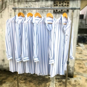 May include: Five long-sleeved button-down shirts hanging on a metal rack. Each shirt is white with vertical blue stripes, featuring a notched collar, a front button placket, and cuffs on the sleeves. The shirts are displayed side by side.