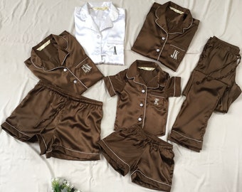 May include: A set of brown satin pajamas with white piping and personalized embroidery. The set includes a short-sleeved top, long pants, and shorts. The initials "A", "JK", "FHK", and "K" are embroidered on the garments.