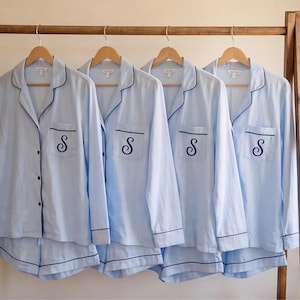 May include: Four light blue pajama sets hanging on wooden hangers. Each set includes a long-sleeved button-down shirt with a notched collar, dark blue piping, and a chest pocket embroidered with a dark blue script letter S, paired with matching shorts.