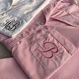 May include: Two sets of pyjamas. One set is white with black embroidered initials on the pocket. The other set is pink and white striped with a pink embroidered initial on the pocket. Both sets have a collared shirt and button closures.