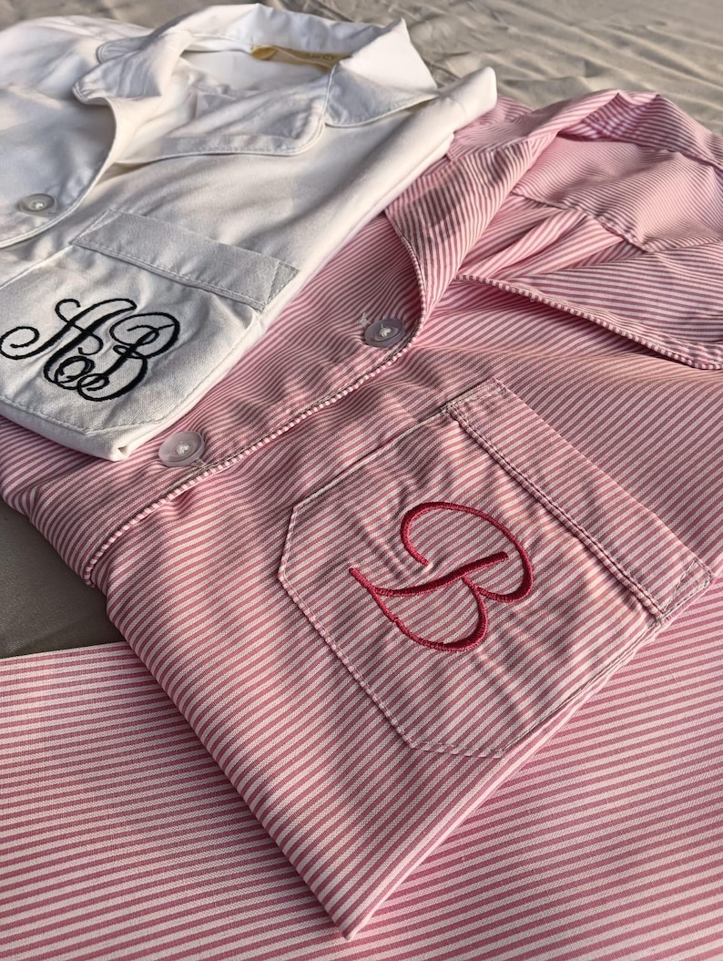 May include: Two sets of pajamas. One set is white with black embroidered initials on the pocket. The other set is pink and white striped with a pink embroidered initial on the pocket. Both sets have a collared shirt and button closures.