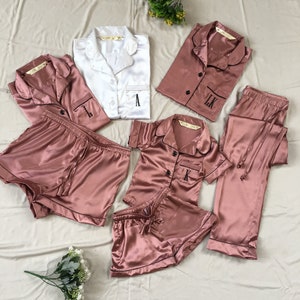 May include: A set of matching pajamas in a dusty rose color with black piping. The set includes a long-sleeved shirt, a short-sleeved shirt, a pair of pants, and two pairs of shorts. Each piece has a single initial embroidered on the front.