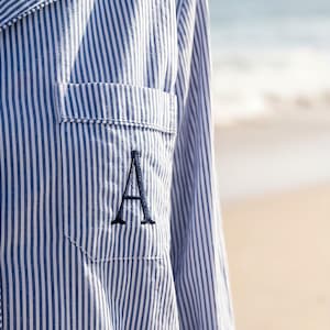 Puede incluir: Camisa de pijama a rayas azules y blancas con un bolsillo en el pecho. El bolsillo presenta una letra "A" bordada en azul oscuro. La camisa tiene mangas largas y una botonadura. El fondo es una escena de playa borrosa.