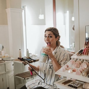 May include: A white button-down pajama shirt with thin blue vertical stripes and a black monogrammed letter M on the pocket. The shirt features a classic collar and long sleeves, worn while holding a disposable beverage cup and a makeup brush.