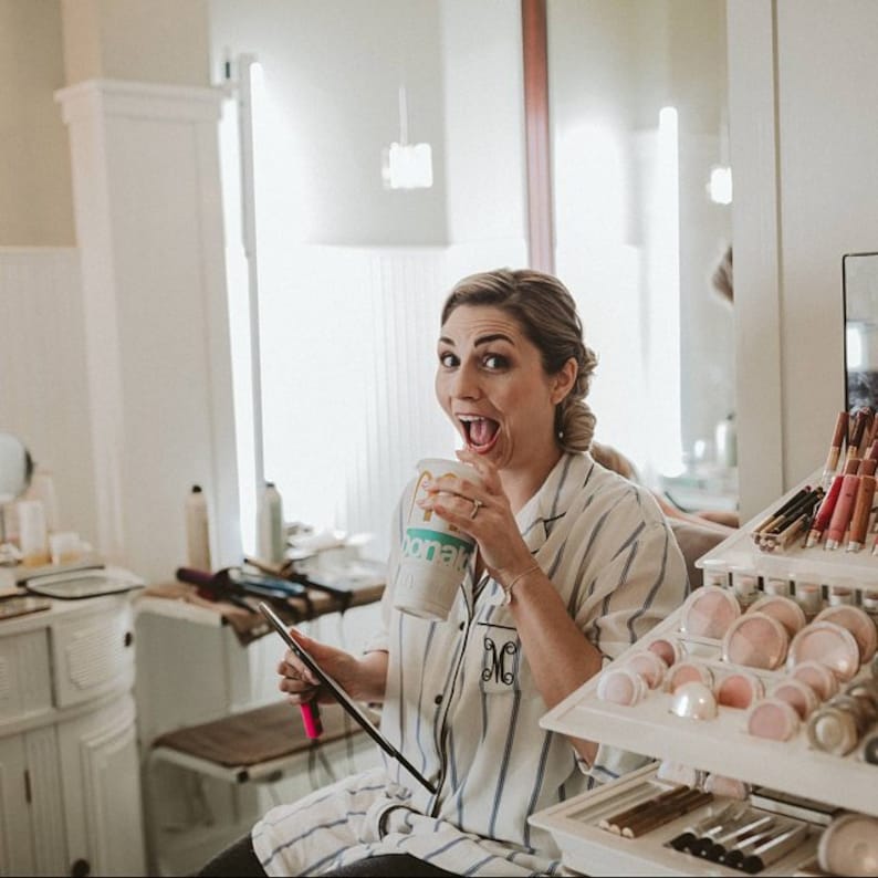 May include: A white button-down pajama shirt with thin blue vertical stripes and a black monogrammed letter M on the pocket. The shirt features a classic collar and long sleeves, worn while holding a disposable beverage cup and a makeup brush.