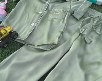 May include: A sage green satin pajama set with a short-sleeved top and long pants. The top has a button-up front and a small pocket with the letter "A" embroidered on it.