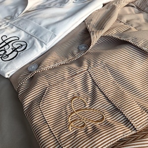 May include: Two button-down shirts, one white and one brown striped, are neatly folded. The white shirt has a black embroidered monogram on the pocket. The brown shirt features a gold embroidered monogram. Both shirts have a classic, tailored design.