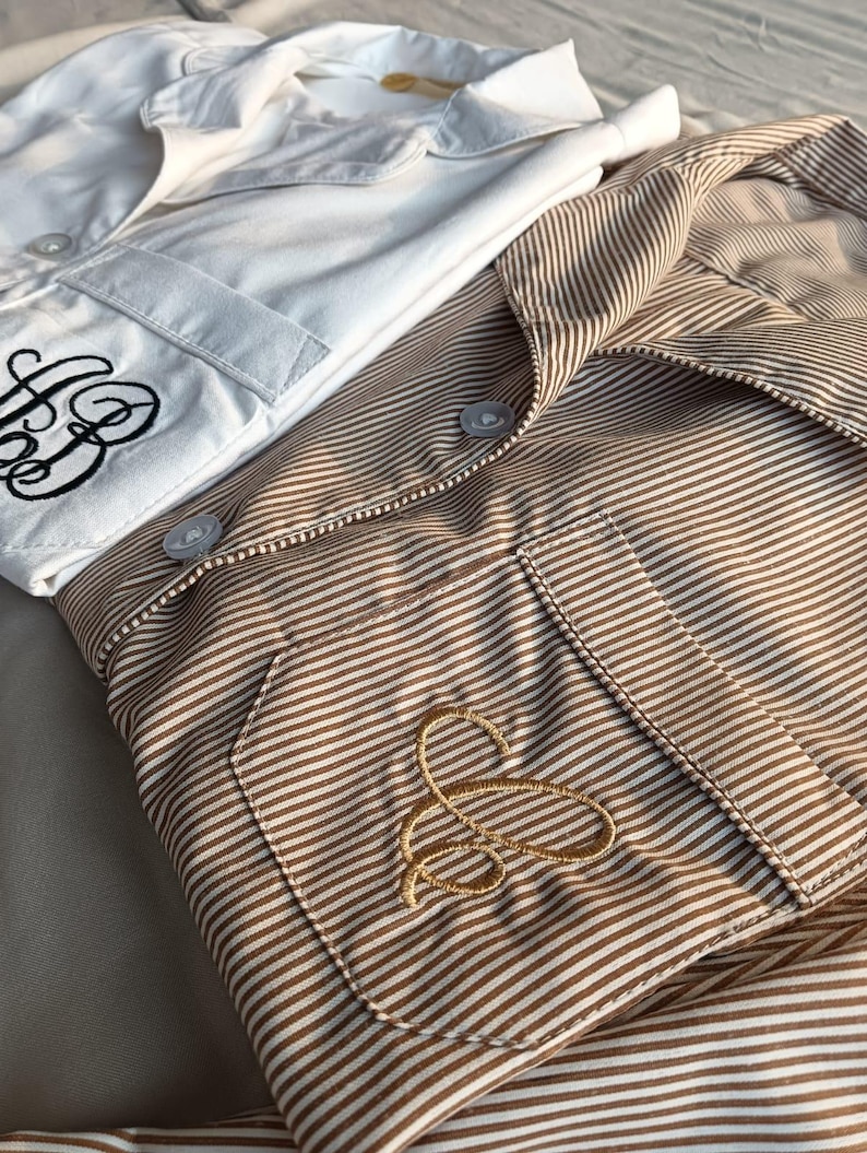 May include: Two button-down shirts, one white and one brown striped, are neatly folded. The white shirt has a black embroidered monogram on the pocket. The brown shirt features a gold embroidered monogram. Both shirts have a classic, tailored design.