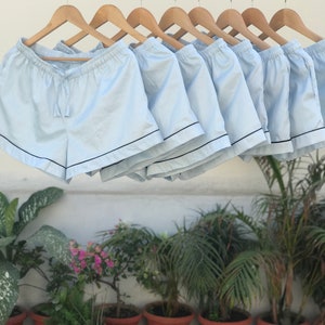 Puede incluir: Una fila de pantalones cortos de pijama azul claro con ribete negro colgados de perchas de madera. Los pantalones cortos est&aacute;n hechos de una tela suave y ligera y tienen una cintura con cord&oacute;n.