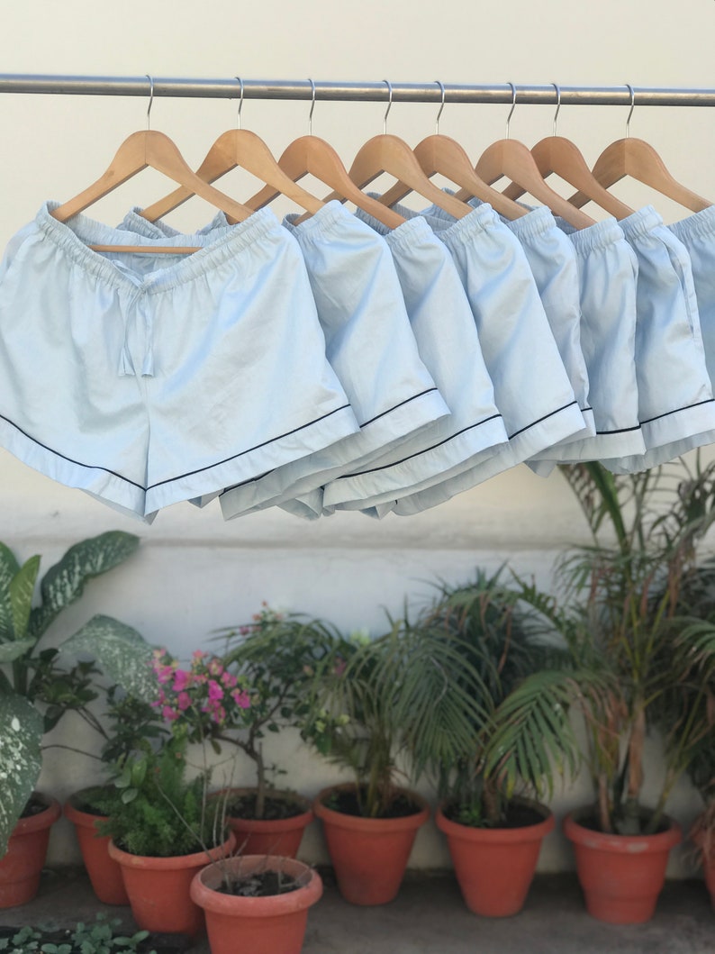 Puede incluir: Una fila de pantalones cortos de pijama azul claro con ribete negro colgados de perchas de madera. Los pantalones cortos est&aacute;n hechos de una tela suave y ligera y tienen una cintura con cord&oacute;n.
