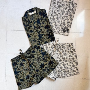 May include: Four-piece pajama set featuring two button-down shirts and two pairs of shorts. One set is olive green with navy floral print, the other is off-white with a black floral pattern. The off-white set has embroidered initials.