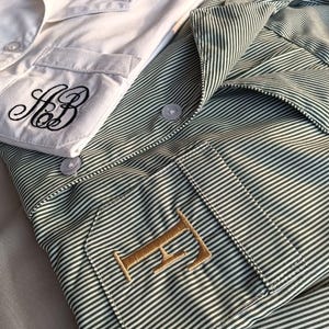 May include: Two pajama tops, one white and one green striped. The white top has a pocket with black embroidered initials. The green striped top has a pocket with a gold embroidered letter. Both tops have button closures and collars.