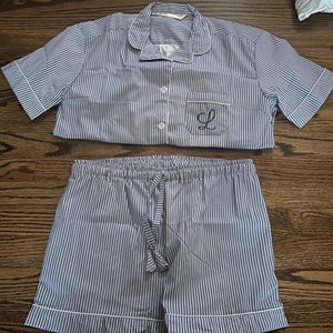 May include: A blue and white striped pyjama set. The short-sleeved top features a notched collar, a breast pocket with an embroidered "L", and button fastenings. The matching shorts have a drawstring waist and white trim.