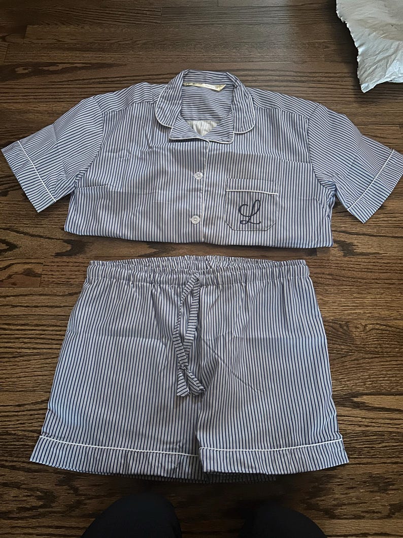 May include: A blue and white striped pyjama set. The short-sleeved top features a notched collar, a breast pocket with an embroidered "L", and button fastenings. The matching shorts have a drawstring waist and white trim.