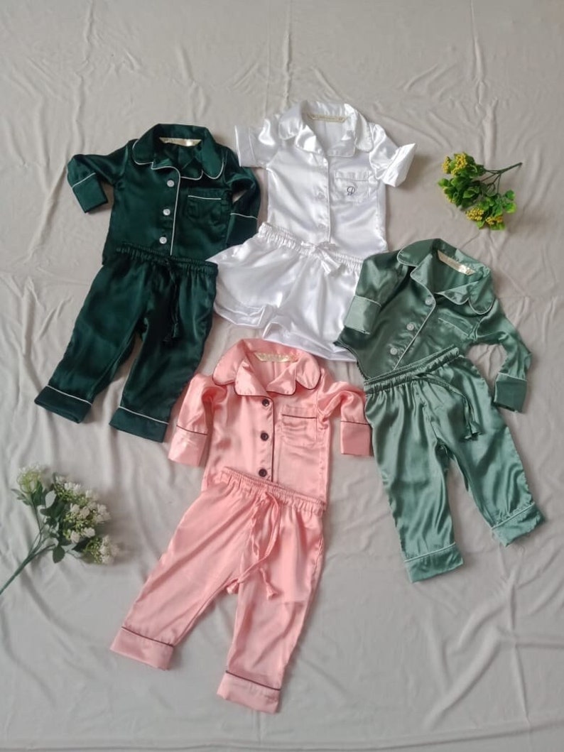 May include: Four sets of pajamas for children, each with a different color top and matching pants. The pajamas are made of a silky material and have a button-up top with a collar. The colors are green, white, pink, and a darker green.