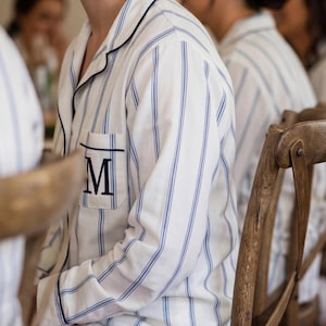 Puede incluir: Camisa de pijama blanca con rayas verticales azules y bolsillo con borde negro con la letra "M". La camisa tiene ribetes negros y la llevan varias personas sentadas en sillas de madera. La escena sugiere una boda o evento similar.