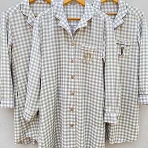 May include: Three gray and white gingham button-down shirts with black trim and personalized initials. The shirts are hanging on wooden hangers.