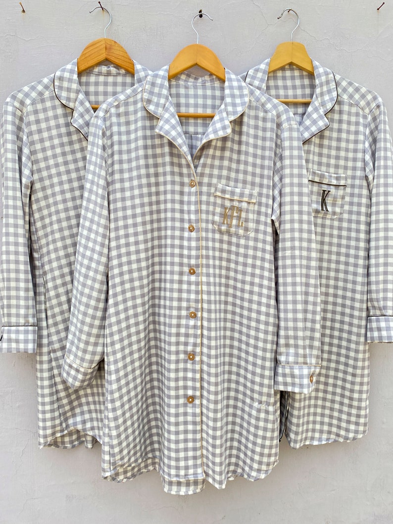 May include: Three gray and white gingham button-down shirts with black trim and personalized initials. The shirts are hanging on wooden hangers.