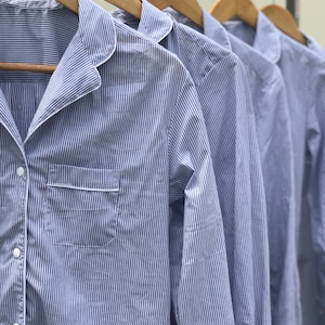 May include: A series of long-sleeved button-down shirts featuring a blue and white pinstripe pattern. Each shirt has a notched collar with white piping, a single chest pocket with white trim, and white buttons, all hanging on wooden hangers.