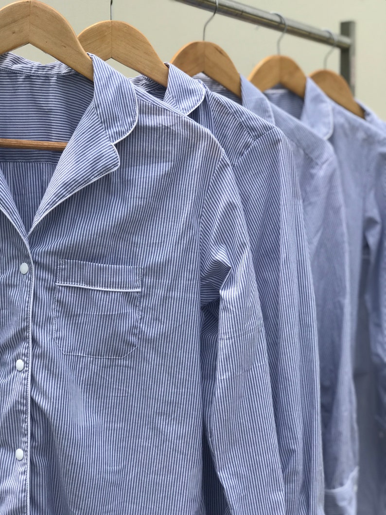 May include: A series of long-sleeved button-down shirts featuring a blue and white pinstripe pattern. Each shirt has a notched collar with white piping, a single chest pocket with white trim, and white buttons, all hanging on wooden hangers.