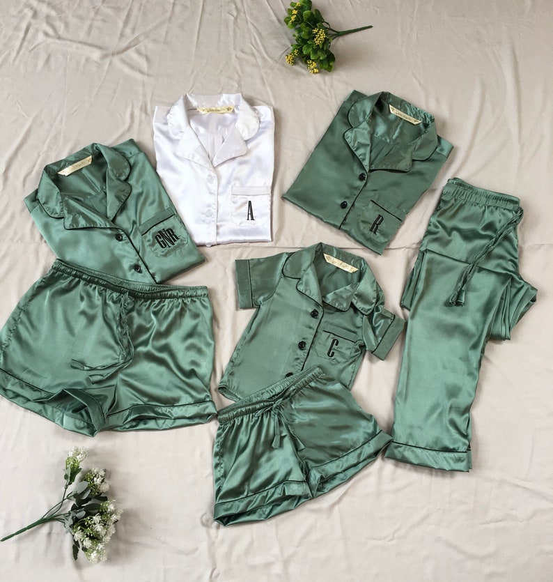 May include: A set of matching pajamas in green and white satin. The pajamas feature a button-up top with a collar and short sleeves. The set includes a pair of shorts and a pair of pants. The tops are personalized with the letters "A", "R", "C", and "GNR".