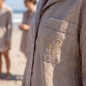 Pode incluir: Close-up de uma camisa de pijama listrada com um bolso e uma letra "C" dourada bordada. A camisa tem uma gola clássica e uma frente abotoada. O fundo mostra duas pessoas em uma praia, sugerindo um tema descontraído.