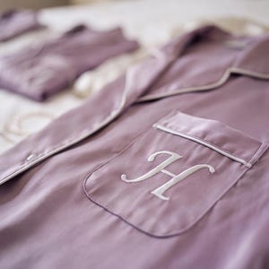 May include: A mauve button-down pajama shirt featuring a white embroidered letter H on the chest pocket. The shirt has a notched collar and white piping trim along the edges of the collar, the front opening, and the top of the chest pocket.