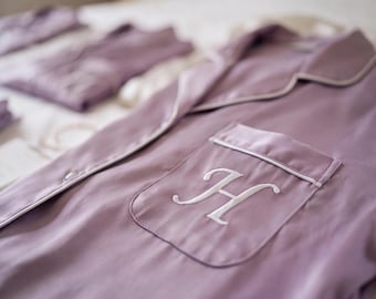 May include: A mauve button-down pajama shirt featuring a white embroidered letter H on the chest pocket. The shirt has a notched collar and white piping trim along the edges of the collar, the front opening, and the top of the chest pocket.