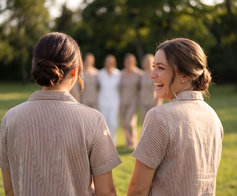 May include: Two people wearing short-sleeved, brown and white striped shirts, standing on a grassy lawn. Other people are in the background, out of focus. The person on the right is smiling. The background is blurred with trees.