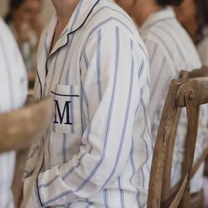 May include: A white long-sleeve button-down pajama top featuring thin vertical blue stripes, a notched collar with dark blue piping, and a single chest pocket embroidered with a large, dark blue capital letter M in a serif font.