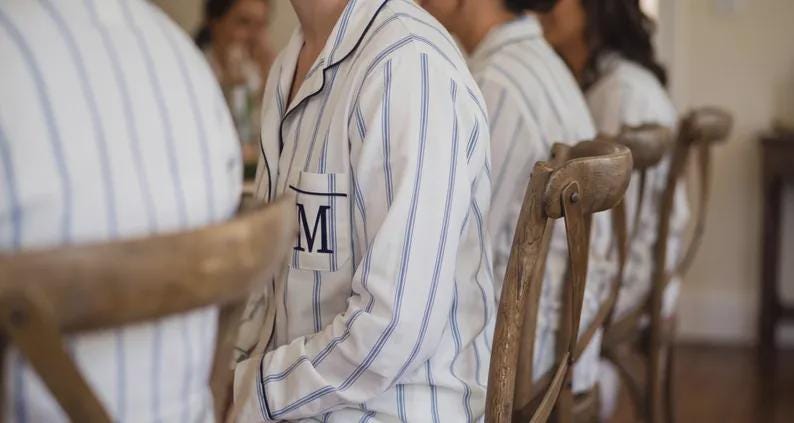 May include: A white long-sleeve button-down pajama top featuring thin vertical blue stripes, a notched collar with dark blue piping, and a single chest pocket embroidered with a large, dark blue capital letter M in a serif font.