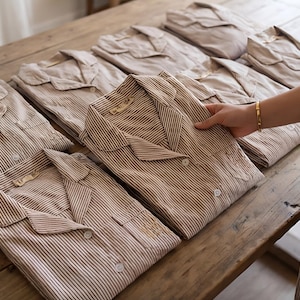 May include: Several beige and brown striped pajama sets are neatly arranged on a wooden surface. One set is being held by a person wearing a gold bracelet. The pajamas have a collared neckline and button-down front.