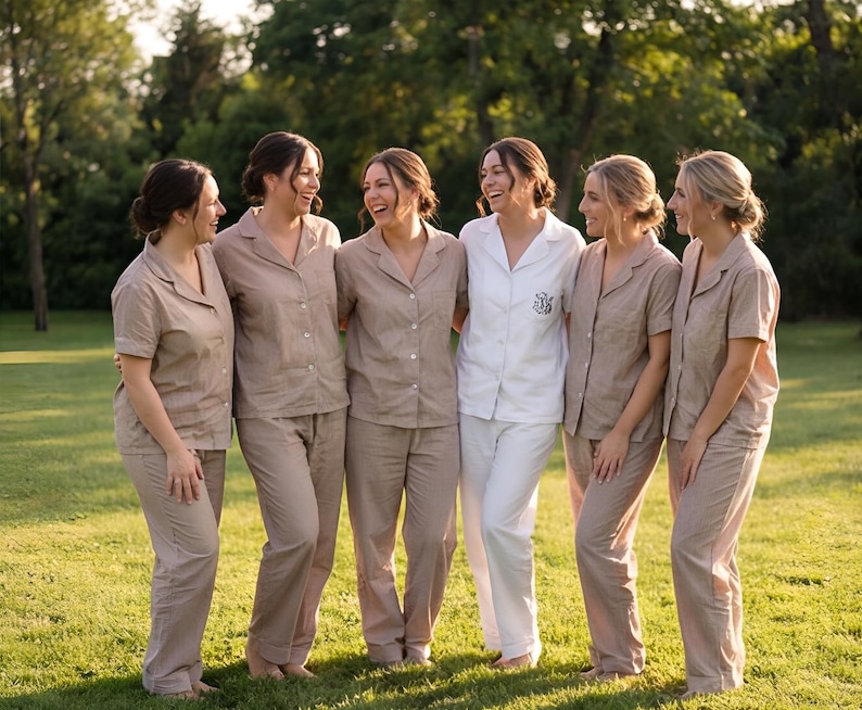 May include: Six people wearing matching pajama sets stand together on a grassy lawn. Five are in tan sets, while one wears a white set. The sets include button-down shirts and pants. The group is smiling and laughing, suggesting a relaxed, social gathering.