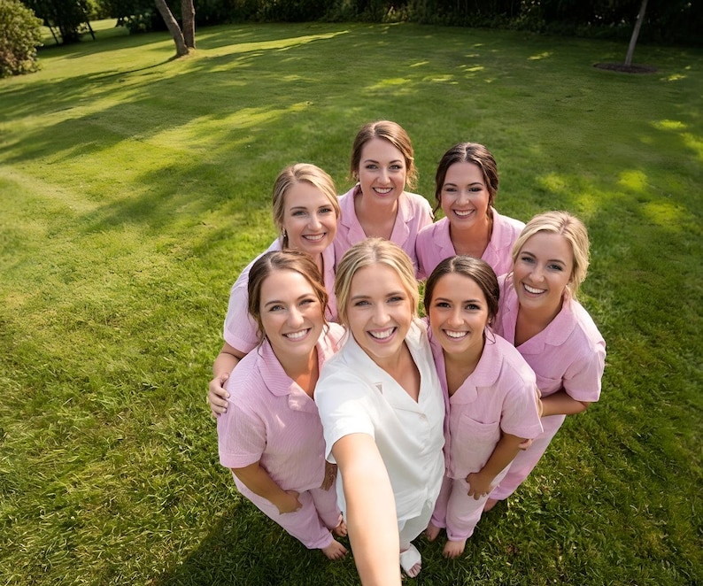 May include: A group of seven smiling people, mostly women, pose for a photo outdoors on a grassy lawn. Most are wearing matching light pink pajama sets. One person is wearing a white robe. The photo is taken from a high angle.