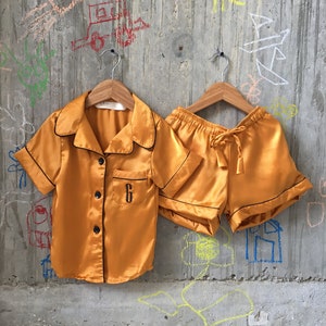 Puede incluir: Un conjunto de pijama de sat&eacute;n amarillo mostaza con ribetes negros y una letra "G" bordada en negro en el bolsillo de la parte superior.