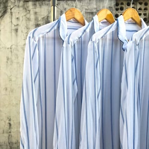 May include: Three long-sleeved shirts made of white fabric with vertical blue stripes. Each shirt features a mandarin collar, a button-down front, and cuffs with a single white button. The shirts are hanging on wooden hangers on a metal rack.