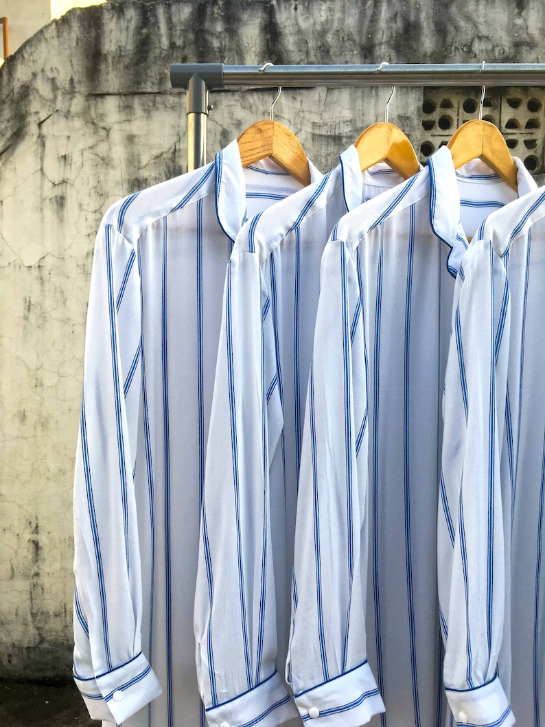 May include: Three long-sleeved shirts made of white fabric with vertical blue stripes. Each shirt features a mandarin collar, a button-down front, and cuffs with a single white button. The shirts are hanging on wooden hangers on a metal rack.