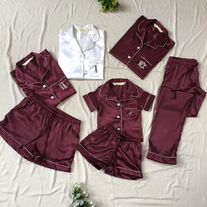 May include: A set of matching burgundy satin pajamas with white piping. The set includes a long-sleeved top, a short-sleeved top, a pair of pants, and two pairs of shorts. Each piece has a white embroidered initial on the front.