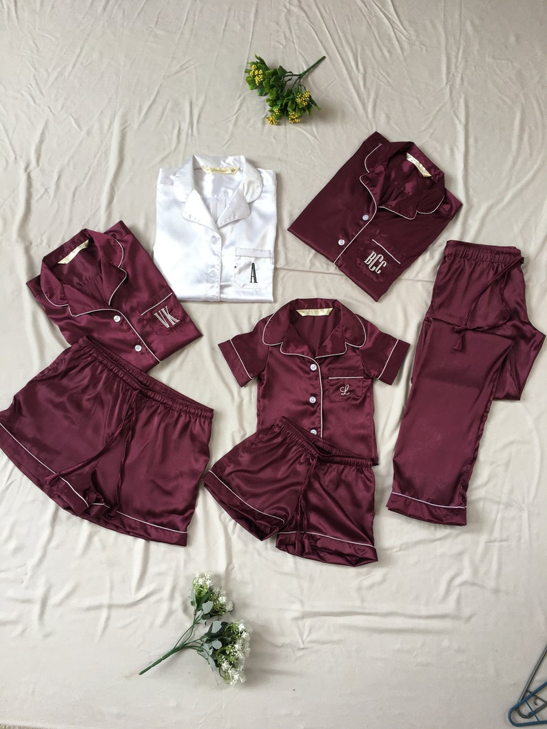 May include: A set of matching burgundy satin pajamas with white piping. The set includes a long-sleeved top, a short-sleeved top, a pair of pants, and two pairs of shorts. Each piece has a white embroidered initial on the front.