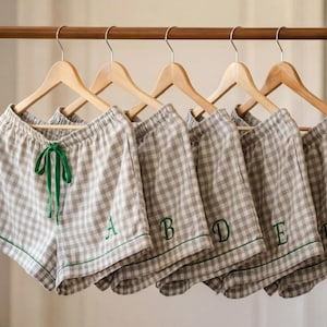 May include: A set of six pairs of gray and white checkered pajama shorts hanging on a wooden rack. Each pair has a green drawstring and trim, with a single embroidered letter. The shorts are on wooden hangers.