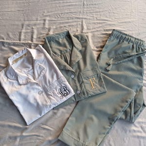 May include: Three sets of pajamas are displayed on a gray surface. One set is white, and the other two are green and white striped. The white set has a monogram on the cuff. The green set has a monogram on the pocket. The pajama pants have a drawstring.